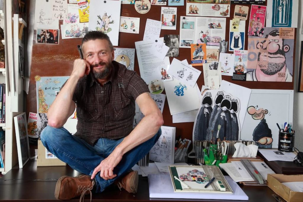 Der Comic-Zeichner Ralf König sitzt im Schneidersitz auf dem Zeichentisch seines Kölner Ateliers.